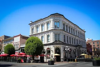 A large white building with many windows sits on the corner of a street. The building looks antique with older style architecture and window shapes. It features a patio with tables and red umbrellas on the sidewalk on the left side.